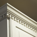 The top crown moulding dentil detail on some creme-color painted solid wood kitchen cabinets. These wall mounted cabinets do not go all the way to the ceiling above them. The lighting is coming from the top right from a ceiling light.